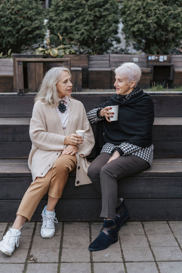 Due amiche anziane che discutono sedute su una panchina e bevono un caffé